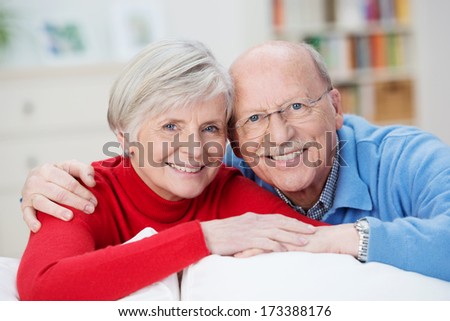 Senior husband and wife smiling happily as they pose together for the camera looking over the back of a comfortable couch in their living room