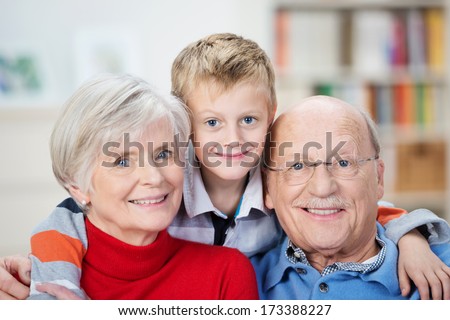 Proud happy elderly grandparents posing with their adorable little grandson hugging them from behind grinning playfully at the camera