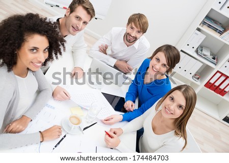 Group of happy multiethnic young businesspeople seated around a table at the office pausing in their meeting to look at the camera