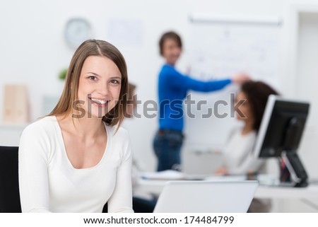 Smiling friendly young businesswoman sitting in the office in front of her co-workers working at their desks in the background