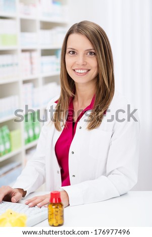 Attractive pharmacist with a lovely smile standing working on the computer behind the counter in the pharmacy