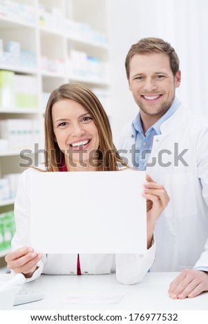 Beautiful pharmacist displaying a blank white sign as she stands behind the counter in the pharmacy with a male colleague
