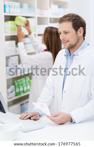 Handsome young male pharmacist checking a prescription on the computer with his female colleague checks supplies behind him