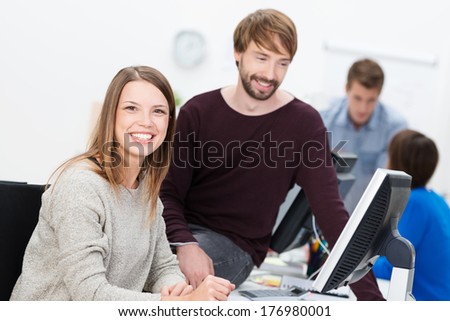Happy business man and woman working together in the office as they share the monitor of a desktop computer with the man sitting comfortably on top of the desk for a better view