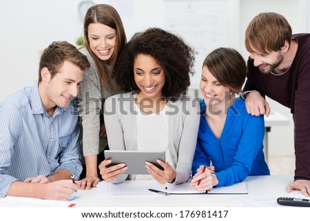 Teamwork at the office with a group of enthusiastic smiling young business people clustered around a pretty African American woman holding a tablet computer