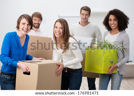 Laughing diverse multiethnic group of young friends moving house carrying cardboard boxes and houseplants in a teamwork concept