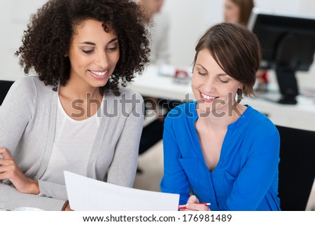 Two smiling businesswomen working on a document as they sit close together at a desk in the office, one is African American