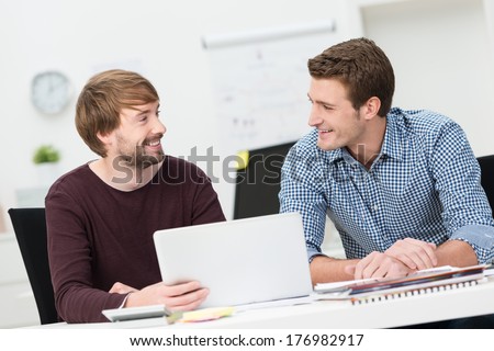 Two Male Friends Working Together In The Office Sitting Smiling And Chatting As They Share A Laptop Computer