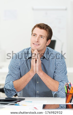 Handsome happy young businessman sitting daydreaming at his desk with a contented smile on his face