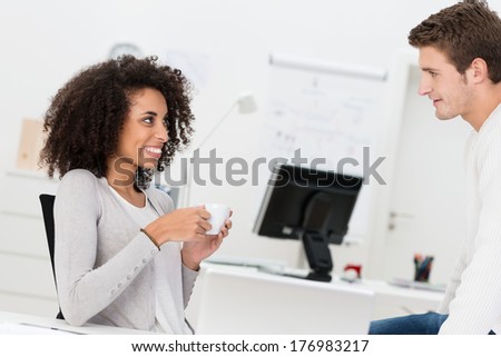 Two business colleagues relax over coffee with a smiling African American woman chatting to a handsome businessman sitting on the desk
