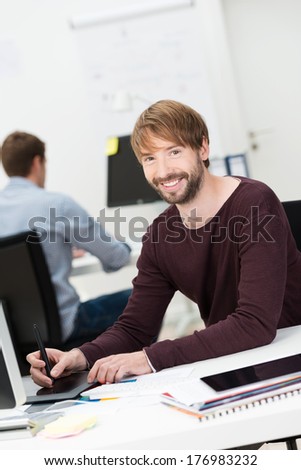 Smiling young man at work in the office sitting working on a computer with a tablet and stylus as his business partner works behind him