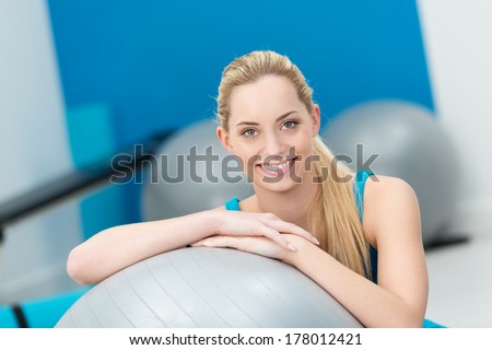 Beautiful friendly woman in a gym pausing to smile at the camera as she rests on a Pilates ball in a health and fitness concept