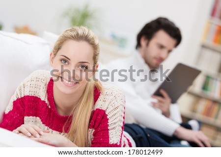 Smiling beautiful young blond woman lying on a sofa relaxing while her husband reads in the background