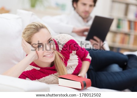 Beautiful young blond woman reading a book as she relaxes on a sofa at home with her husband in the background