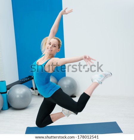 Beautiful happy woman leaping in a gym in a graceful pose with her knees bent and arms outstretched against a backdrop of pilates gym balls