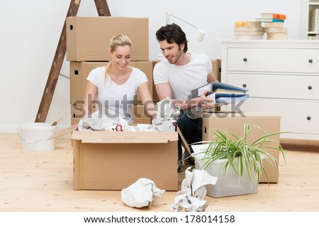 Happy Young Couple Unpacking In Their New Home Kneeling On A Bare Wooden Floor Unwrapping Items From A Large Brown Cardboard Box