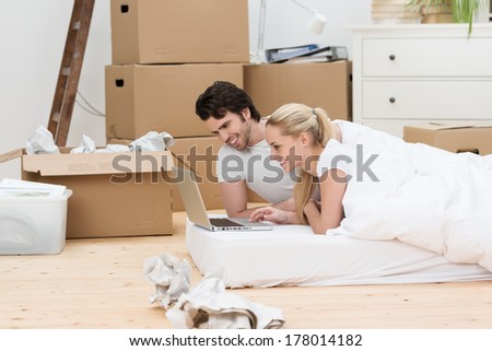 Happy couple taking a break from moving lying on a mattress on the wooden floor in their new house checking their email on a laptop surrounded by cardboard boxes