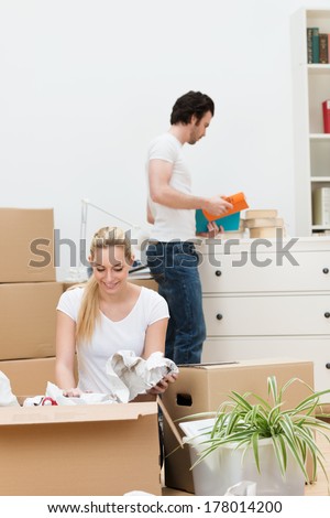 Attractive young blond woman kneeling on the floor unwrapping items from a packing carton in their new home as her husband puts everything away in a teamwork concept