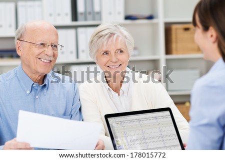 Smiling senior couple meeting with a broker in her office sitting close together holding paperwork and listening to her speak