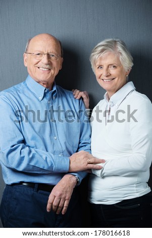 Happy confident elderly couple posing holding hands tenderly smiling at the camera against a dark grey background
