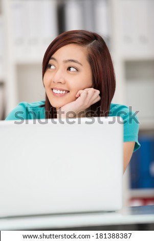 Pretty smiling Asian student sitting thinking with her chin on her hands as she sits behind her laptop staring off to the side daydreaming