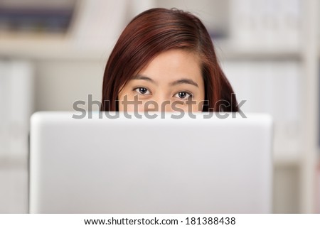 Young Asian woman working at a computer peering over the top of the screen at the camera with just her eyes visible