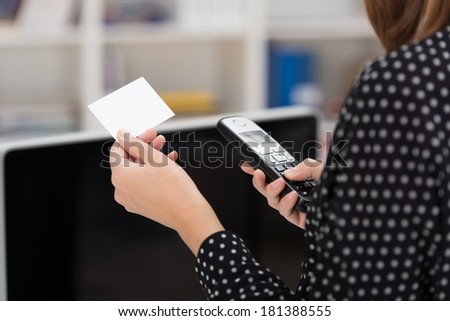 Woman dialing out on her mobile phone using contact details from a blank white business card with copyspace that she is holding in her hand