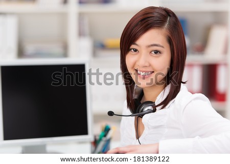 Smiling young Asian woman with a headset around her neck sitting at her desk in the office in front of a desktop computer taking a break from her call centre or reception duties to smile at the camera