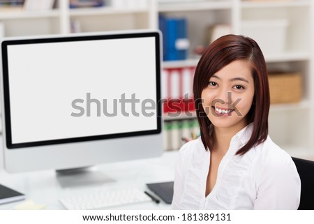 Attractive young Asian businesswoman using a desktop computer sitting in front of the blank monitor turning to smile at the camera