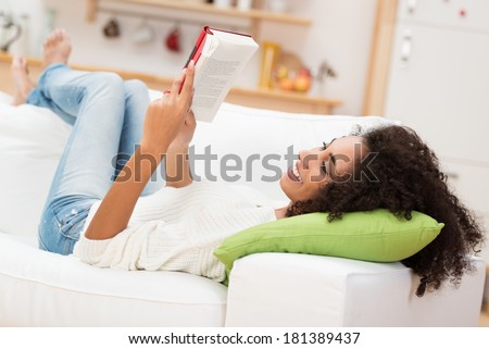 Beautiful young African American woman reading a book lying comfortably on her back on a couch in the living room in her jeans