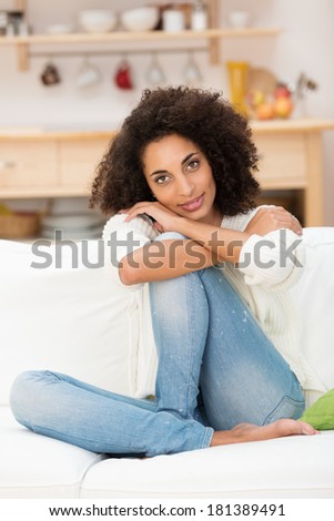 Beautiful young African American woman in a pensive mood sitting barefoot in jeans on her couch in the living room at home
