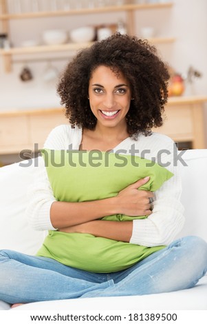 Beautiful African american woman hugging a pillow as she sits cross legged in her jeans on a sofa in the living room smiling at the camera