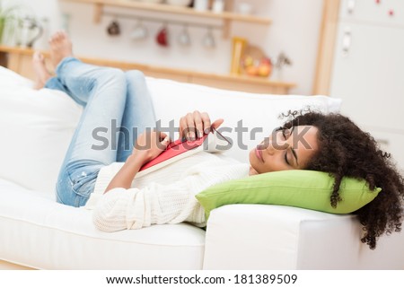 Tired attractive young African American woman fallen asleep with her book on her stomach as she relaxes on her back on a sofa in the living room