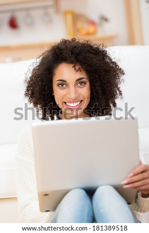 Beautiful young African American student sitting on the floor in her living room with her laptop on her knees looking at the camera with a lovely friendly smile