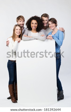 Happy friendly group of multiethnic young friends standing grouped behind a blank white sign that they are holding in the vertical position with copyspace for your text