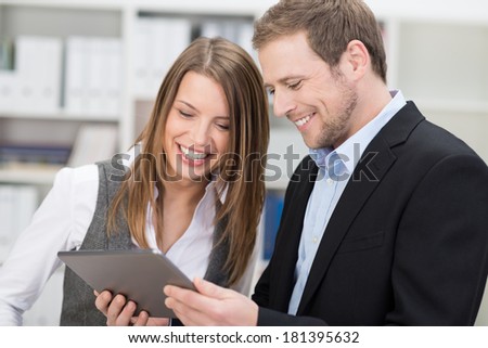Two attractive stylish young co-workers smiling at information on a tablet held by the man as they stand together in the office