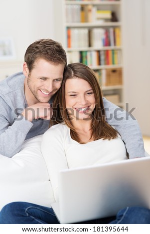 Attractive affectionate young couple relaxing together on a sofa at home grinning in amusement at content on the laptop screen