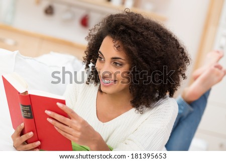 Beautiful Afro-American woman reading a book lying barefoot on a sofa smiling with pleasure as she reads