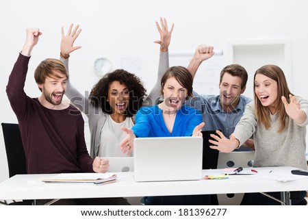 Excited successful business team of diverse multiethnic young people sitting at a table in the office cheering exuberantly as they celebrate a successful outcome on the laptop computer