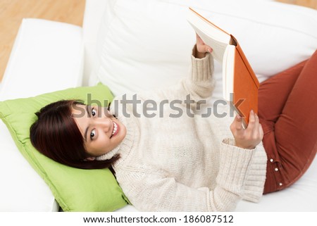 Happy young Asian student studying at home lying on her back on a comfortable sofa with a book smiling up at the camera