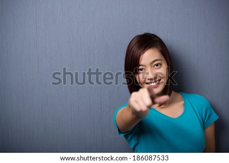 Vivacious smiling young Asian woman pointing at the camera with her finger as she stands against a dark grey background with vignetting and copyspace, focus to her face