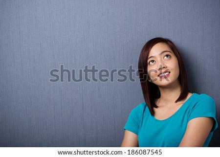 Young Asian woman lost in thought standing against a dark background with copyspace looking up into the air with a smile