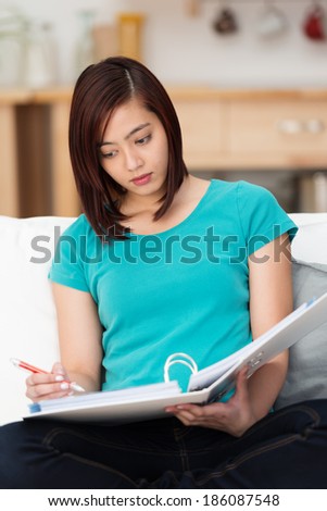 Attractive young Asian student studying at home sitting cross legged on a sofa with a file of notes on her lap