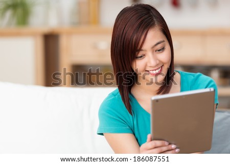 Beautiful young Asian woman browsing the internet on a handheld tablet computer as she relaxes on a sofa at home