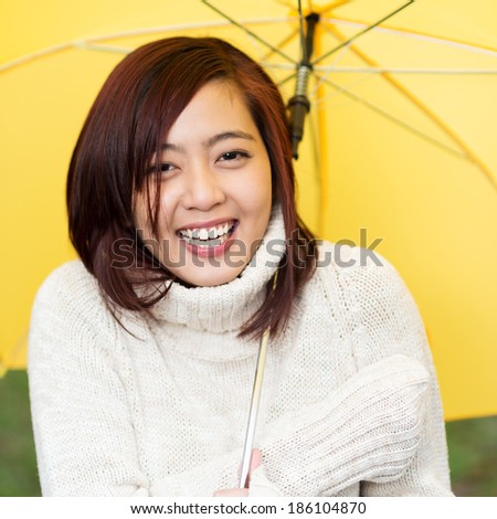 Beautiful happy young Asian woman sheltering under a yellow umbrella in a warm polo neck sweater standing laughing at the camera