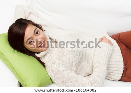 Young Asian woman having a lazy day lying with her head on a cushion on a comfortable sofa looking up with a smile