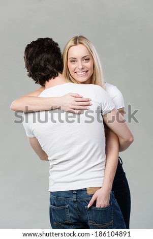 Beautiful blond woman giving her husband a hug looking over his shoulder to smile at the camera, on grey
