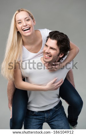 Young couple having fun riding piggy back laughing merrily as the wife rides on her husbands back, on grey