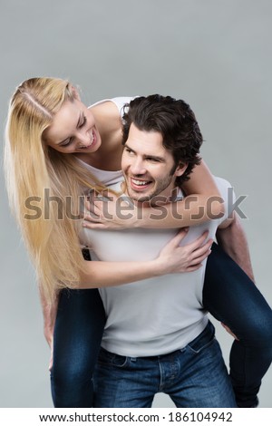 Playful couple riding piggy back together laughing as they young woman with long blond hair rides on her husbands back, on grey