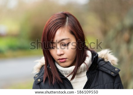 Thoughtful young Asian woman in a warm polo-neck standing outdoors looking down at the ground with a sad expression